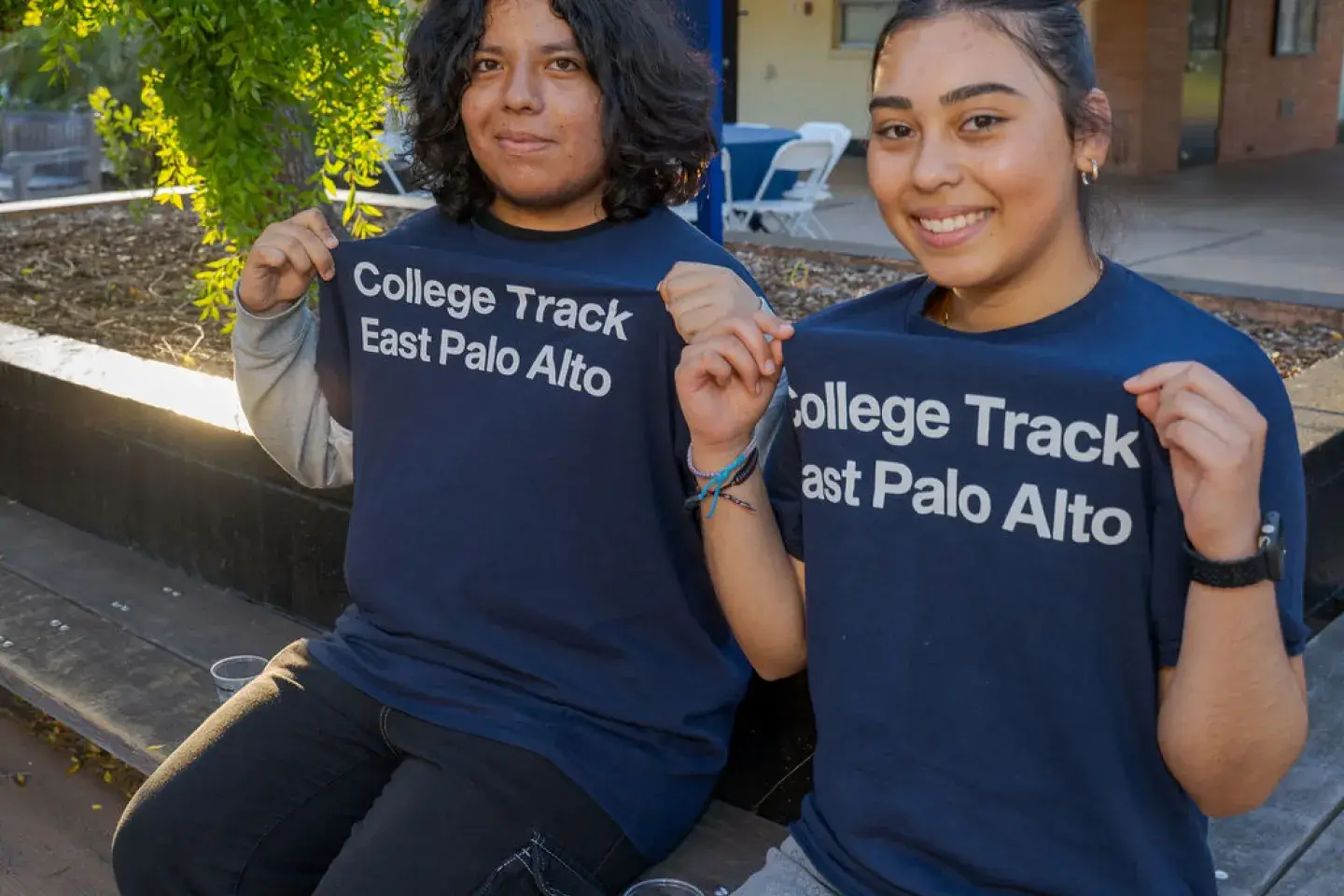 Two students seated outside holding up their matching shirts that read, "College Track East Palo Alto"
