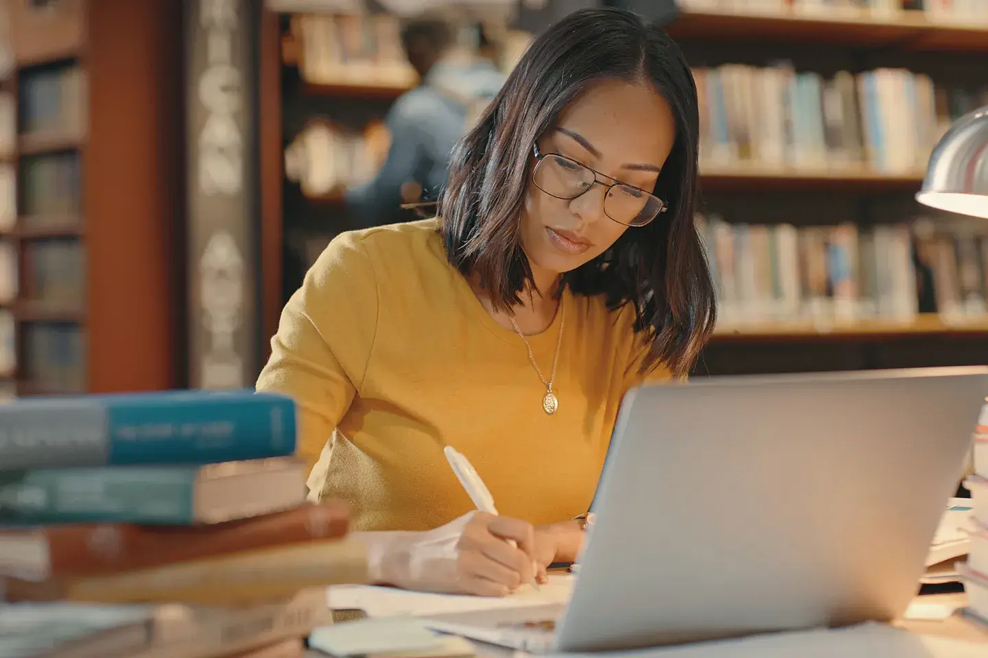 A young woman in a yellow shirt working in the library on her computer with a small stack of books next to her