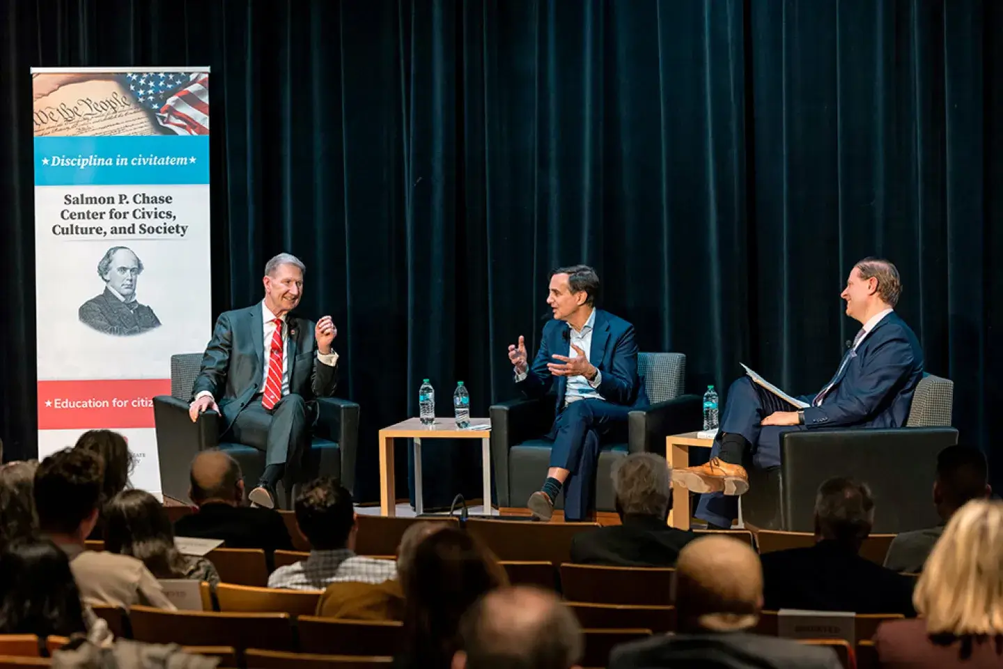 Senior university officials sit in conversation on a stage in front of a small audience of onlookers.