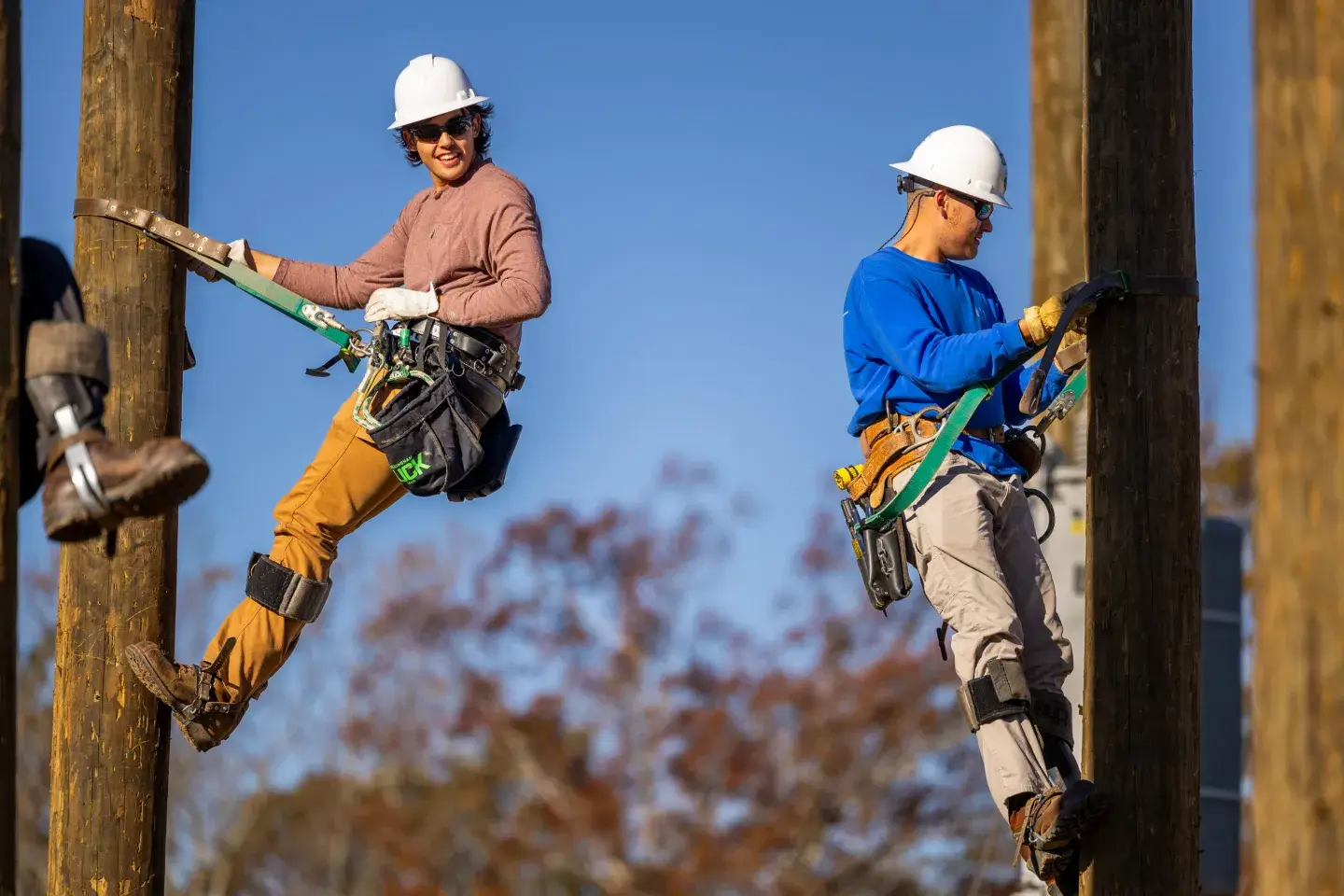 two young men wearing hardhats suspended on logging poles in climbing gear 