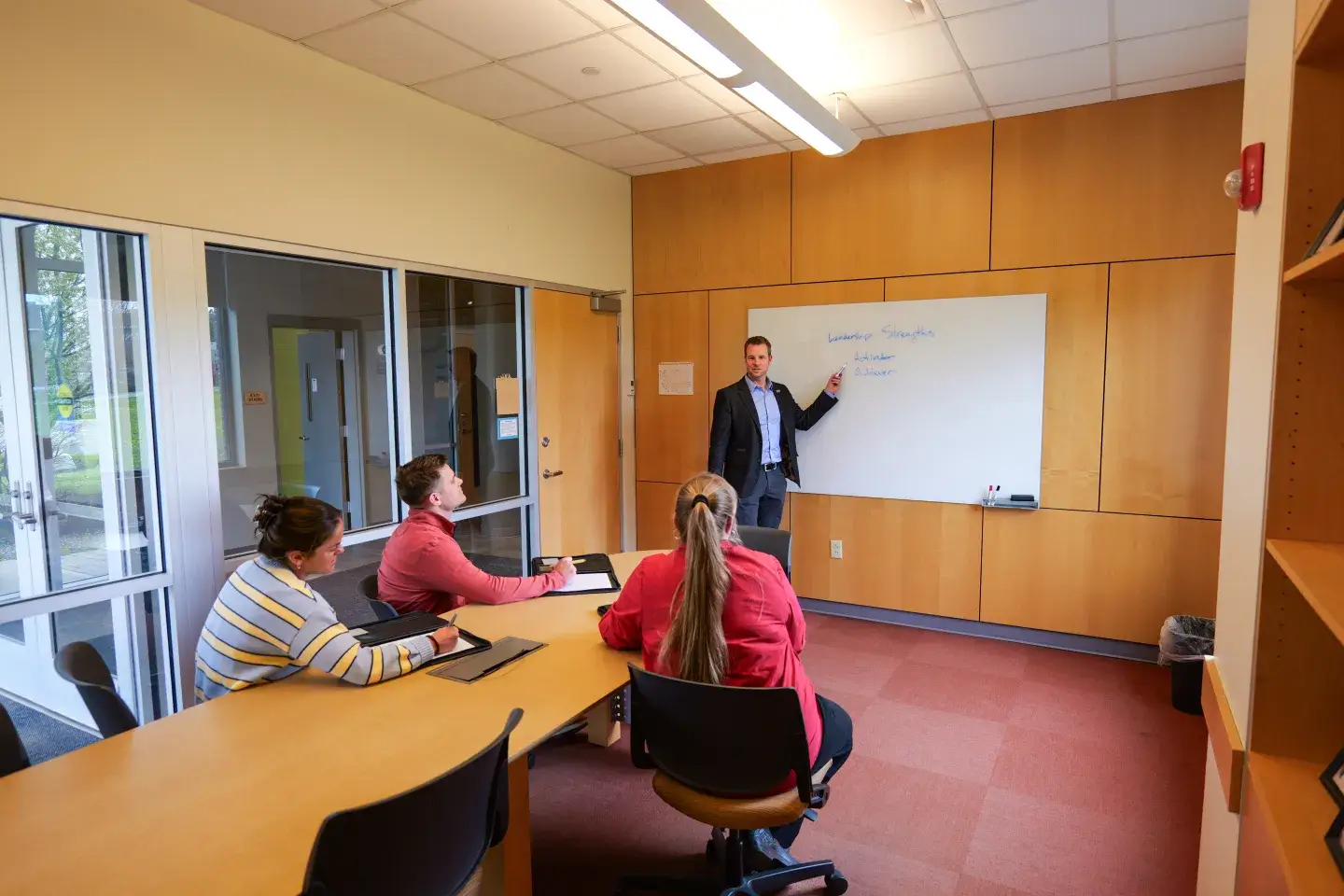 An instructor wearing business casual clothing stands in front of a whiteboard in a classroom with three adult students sitting around a table taking notes.