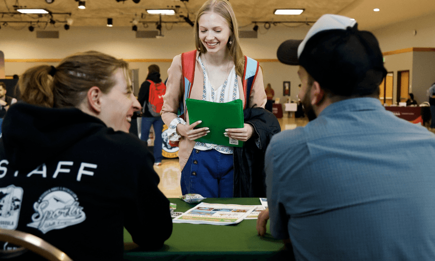 A student with a folder talking to two older students at a resource fair behind a table