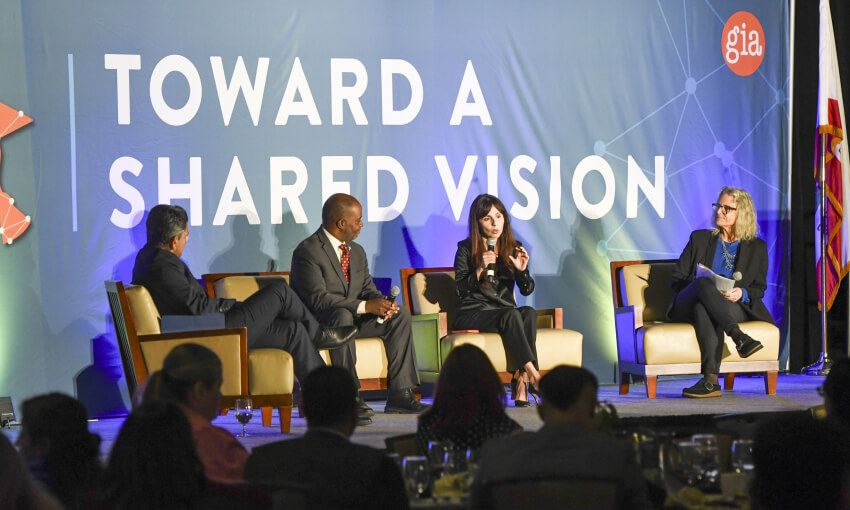 Panelists on stage at a conference with a sign behind them that reads "toward a shared vision"