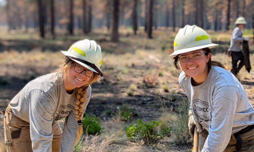 Two young women with hard hats and protective glasses in the woods holding long tools