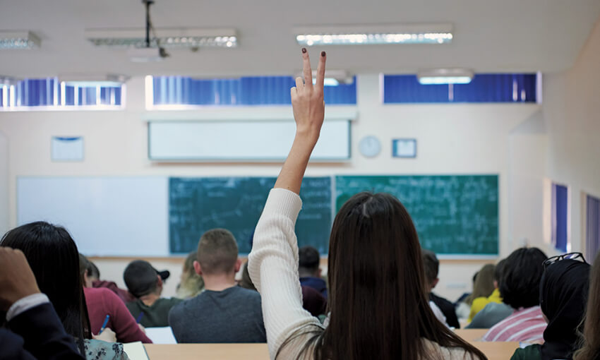 A view from the back of high school students facing a blackboard in a classroom. A student in the center of the class raises their hand.