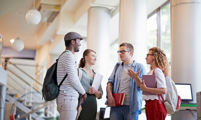 Students are seen standing in a university building engaged in casual conversation while holding their schoolbooks and belongings.