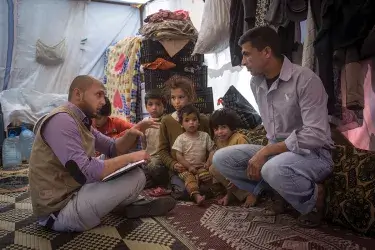 Two adults sit on a rug in a makeshift tent surrounded by crates, clothing, and large plastic water jugs. Five children sit beside them. One of the adults has a notebook and gestures as if explaining something to the other. The other adult faces him attentively.