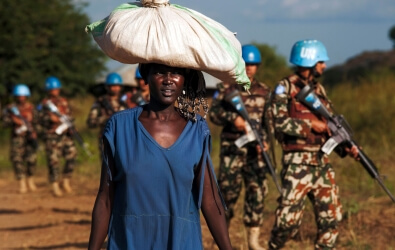 African woman carries bag on her head while UN peacekeepers patrol the background