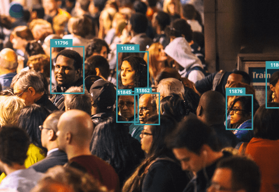 A crowded subway platform shows several commuters turning to face the viewer. Their heads are surrounded by digital boxes that label each an arbitrary number, implying categorization by facial recognition software. The commuters appear to be of various races and ages.