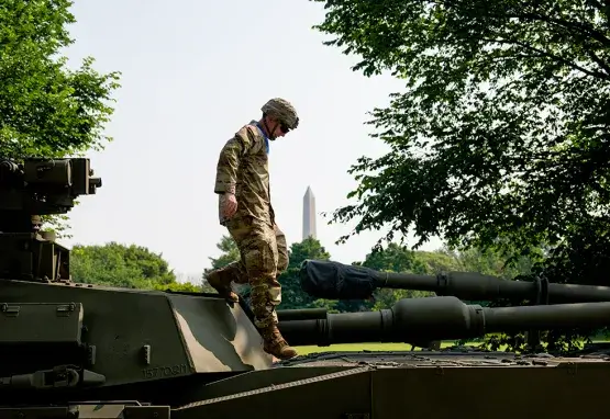 A U.S. Army soldier in uniform and headgear steps on top of an armored fighting vehicle. Behind him are trees, a clear blue sky, and a view of the Washington Monument in Washington, D.C.