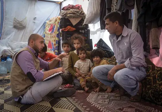 Two adults sit on a rug in a makeshift tent surrounded by crates, clothing, and large plastic water jugs. Five children sit beside them. One of the adults has a notebook and gestures as if explaining something to the other. The other adult faces him attentively.