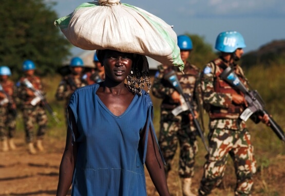 African woman carries bag on her head while UN peacekeepers patrol the background