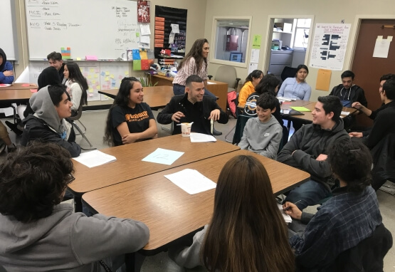 Candid photograph of a teacher sitting around a table and laughing with several high school English students