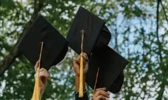 Three hands holding tasseled mortarboards against trees and sky.