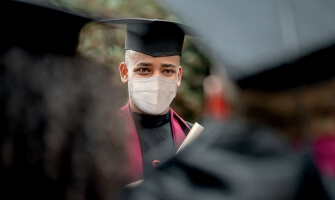 A person with light brown skin and a shaved head wears a graduation cap and gown, as well as a surgical mask. They face their fellow graduates. 