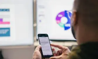 Data on screens in background with man using phone in the foreground. 