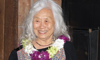 A photo of Maxine Hong Kingston, a person with brown skin and long wavy white hair. She wears a black dress under a multicolored scarf, and a necklace of white, green, and purple flowers. She looks to her right and smiles.