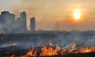 Fire raging in foreground with buildings in the background and sun.