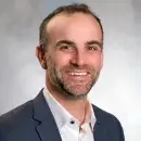 A photo of Daniel Barron, a person with light skin and short brown hair, wearing a dark business suit and white shirt and smiling at the viewer.