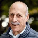 A photo of Richard G. Frank, a person with light skin and short gray hair, wearing a purple jacket and white shirt, and smiling at the viewer.