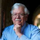 A photo of Henry T. Greely, a person with light skin, gray hair and gray mustache, wearing glasses and a blue shirt, facing the viewer with his hand on his chin.