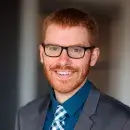 A photo of Nicholas Jacobson, a person with red hair and a red beard and mustache, wearing glasses and a gray suit and blue shirt,  and smiling at the viewer.