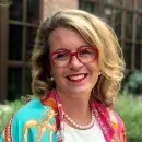A photo of Kacie Kelly, a person with light skin and long blonde hair, wearing red glasses and colorful business attire, and smiling at the viewer.