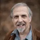 A photo of Arthur Kleinman, a person with light skin, gray hair, and a gray beard and mustache, wearing a brown jacket and blue shirt, and smiling at the viewer.