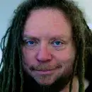 A photo of Jaron Lanier, a person with light skin and long brown hair, and smiling at the viewer.