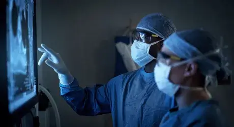 Two medical professionals wearing surgical scrubs, masks, and protective eyewear are examining a medical scan on a screen in a dimly lit room.