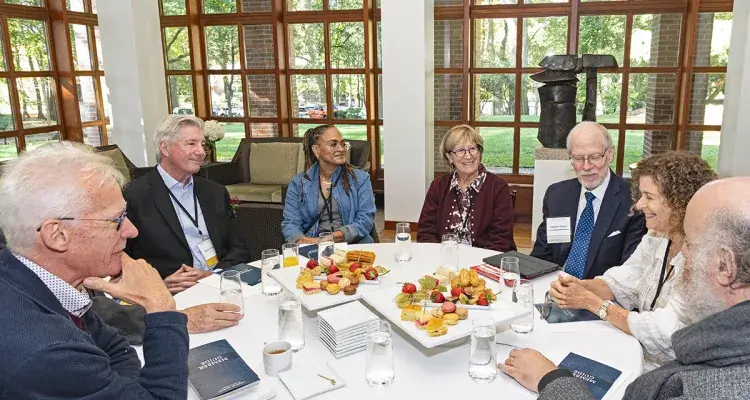 Seven professionally dressed adults in conversation seated at a table in a well-lit room.