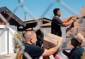 Three people load boxes onto a truck. Two are holding one box, while another person is reaching for the next box. 