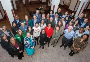 A group of 37 adults in business casual attire pose together for a photo taken from an elevated angle with everyone smiling up at the camera. The subjects in the photo are standing in a space with tiled flooring and white columns with wooden trim. 