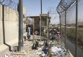 A cement expanse with a gravel floor is fenced and surrounded by barbed wire in Afghanistan. Brown people crowd the other side of the fence and face away from the camera.