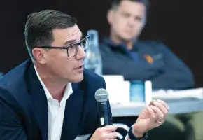 A person in business attire speaks into a microphone and another person in business attire looks forward.