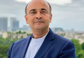 A headshot of Don M. Randel Award recipient Anthony Appiah, a person with short, dark hair and brown skin. Appiah wears a dark blue blazer and white dress shirt, with a blurry skyline view behind him. 