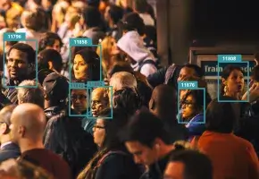 A crowded subway platform shows several commuters turning to face the viewer. Their heads are surrounded by digital boxes that label each an arbitrary number, implying categorization by facial recognition software. The commuters appear to be of various races and ages.