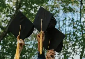 Three hands holding tasseled mortarboards against trees and sky.
