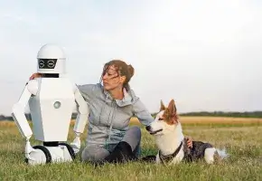 A person sitting in a field with their arms around a dog on their right and a robot on their left.