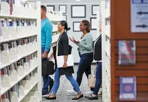 Four people walk through the stacks of a library, two are fully visible at the end of an aisle. One has brown skin and long black hair. The other has brown skin and long light brown hair. Both are smiling.