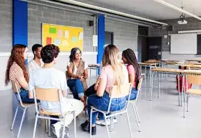 Seven people sitting in chairs in a circle in a classroom setting. Their attention is focused on one member of the group, who is speaking. 