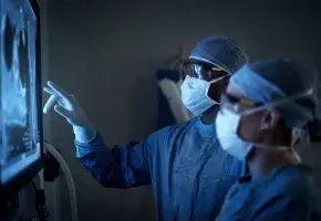 Two medical professionals wearing surgical scrubs, masks, and protective eyewear are examining a medical scan on a screen in a dimly lit room.