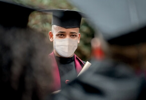 A person with light brown skin and a shaved head wears a graduation cap and gown, as well as a surgical mask. They face their fellow graduates. 