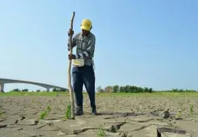 A man in a hard hat standing on parched earth is monitoring drought conditions.