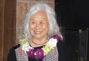 A photo of Maxine Hong Kingston, a person with brown skin and long wavy white hair. She wears a black dress under a multicolored scarf, and a necklace of white, green, and purple flowers. She looks to her right and smiles.