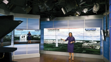 A photo of a television studio shows a camera in the foreground and a meteorologist in the background gesturing toward a monitor where a landscape view of Washington D.C. is displayed. The meteorologist has straight brown hair and brown skin, and wears a purple dress and brown heels. A smaller monitor is a few feet away, which shows a weather forecast along the bottom of the screen. The rest of the screen mirrors the image of the meteorologist.