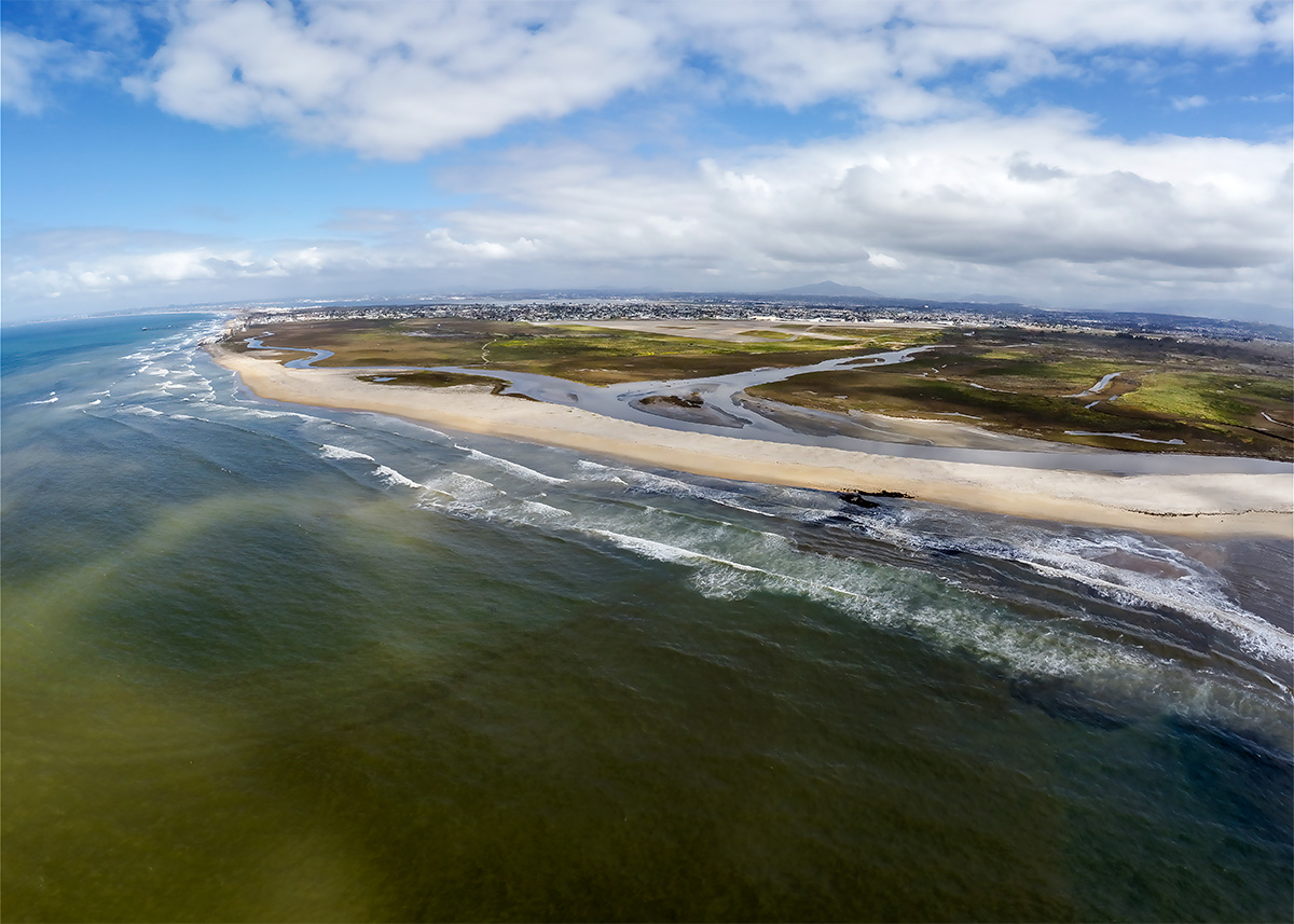 Aerial view of the Tijuana River, featuring a sandy beach where the river meets the ocean. Waves gently crash onto the shore. The background shows green wetlands with winding river paths.