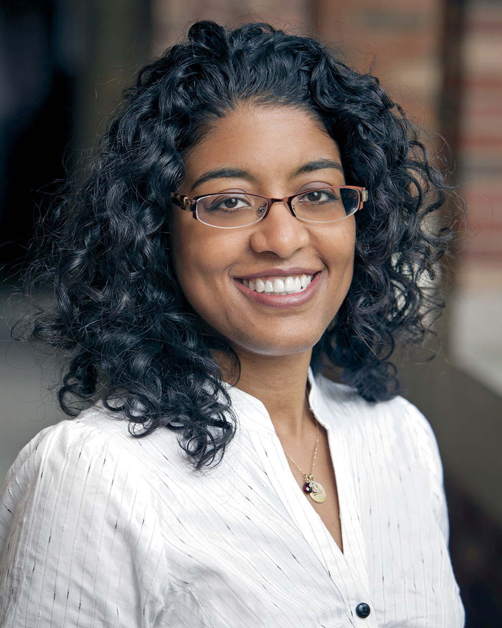 A headshot of Tracey L. Meares wearing wire glasses, a gold necklace, and a white and silver shirt. Meares has brown skin, brown eyes, and long curly black hair. She faces the viewer and smiles. 