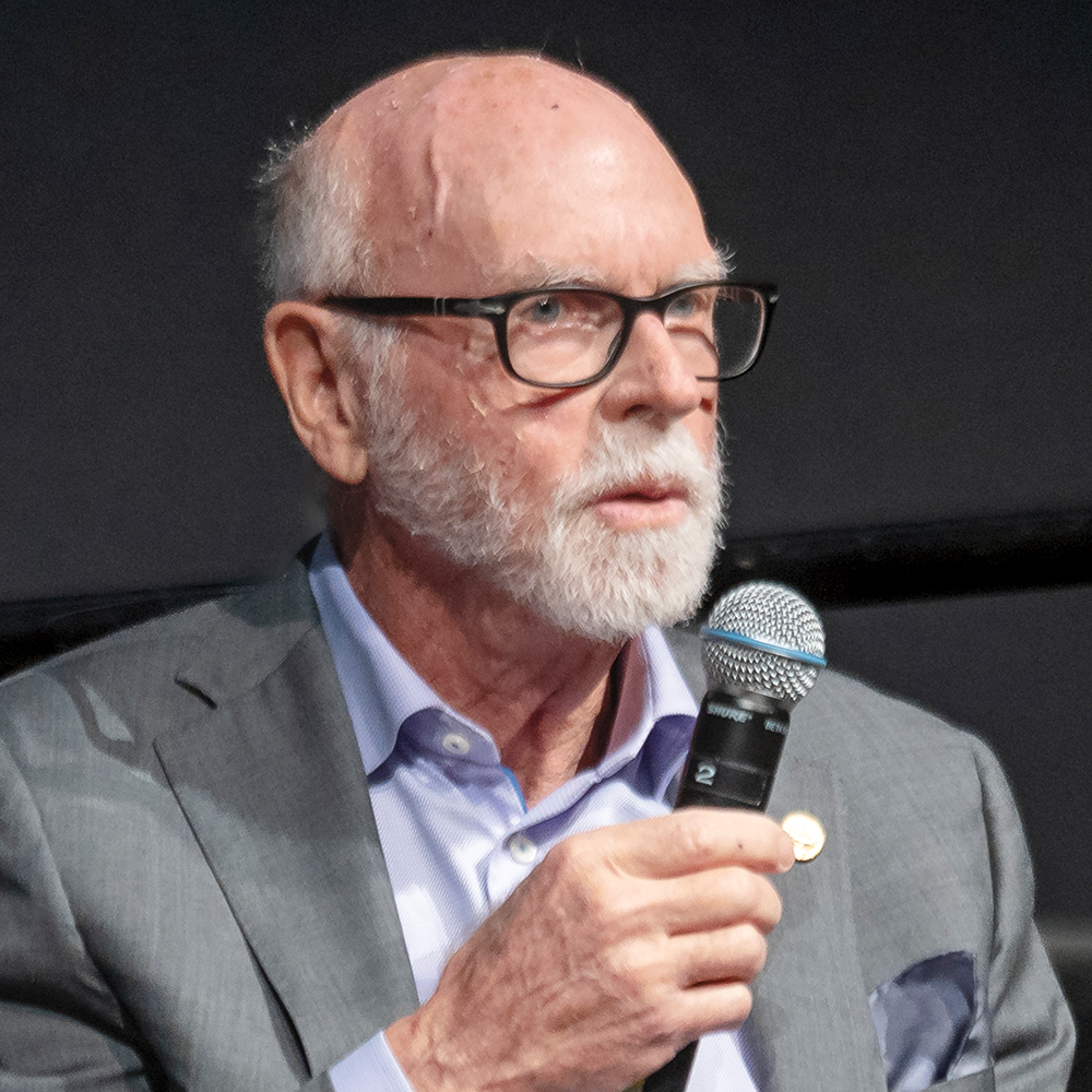 A man with white hair and a white beard and mustache, wearing glasses and a business shirt and jacket, is speaking into a microphone that he is holding in his hand.