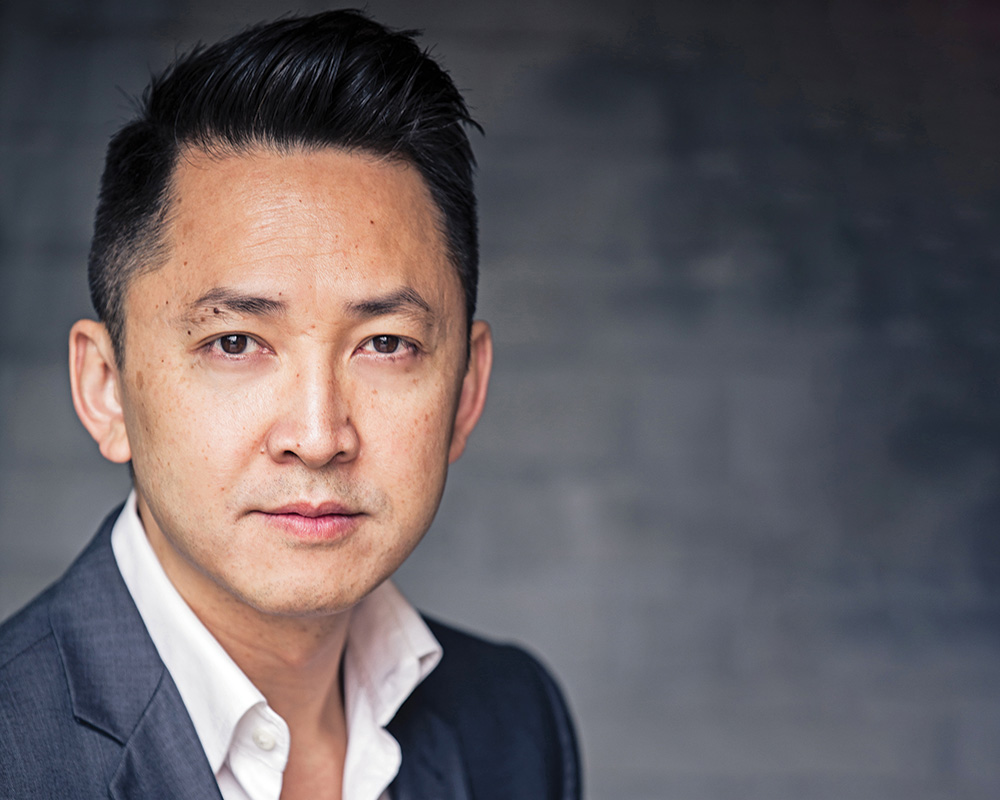 A photo of Viet Thanh Nguyen, a person with brown skin and short straight black hair. He wears a white collared shirt and a gray suit. He faces the camera.